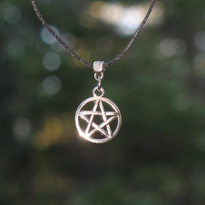 Puede incluir: Collar con colgante plateado con un diseño de pentagrama dentro de un círculo. El colgante cuelga de un cordón fino y oscuro. El fondo es una vista borrosa de follaje verde.