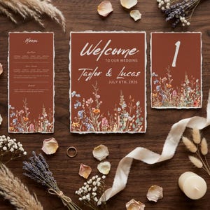 Puede incluir: Un conjunto de papelería de boda que incluye un menú, un cartel de bienvenida y una tarjeta de número de mesa con bordes irregulares. Las tarjetas son de color terracota con texto blanco y una ilustración de flores silvestres. El cartel dice Welcome to our wedding Taylor & Lucas July 6th, 2026.