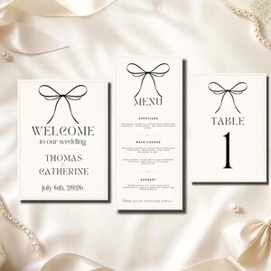 Puede incluir: Tres tarjetas de papelería de boda en color blanco roto con un gráfico de lazo de línea negra en la parte superior. La tarjeta izquierda dice Welcome to our wedding, Thomas & Catherine, July 6th, 2026. El menú central lista platos y la derecha Table 1.