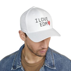 Könnte beinhalten: Weiße Baseballkappe mit dem Text "I LOVE EDM" in grauen Buchstaben und einem roten Herz. Die Kappe besteht aus einem glatten Stoff und hat einen gebogenen Schirm. Die Kappe wird von einer Person mit einer Jeansjacke getragen.