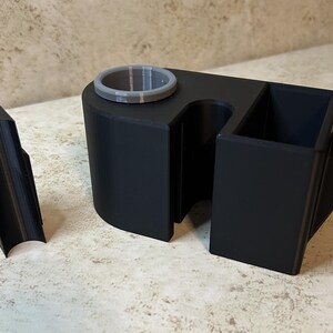 May include: A black 3D-printed car console organizer with a cup holder, a rectangular storage compartment, and a phone holder. A separate black piece is also included. The organizer is designed to fit in a car.