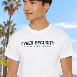 May include: A white short-sleeved t-shirt featuring the text CYBER SECURITY in a black pixelated font, with the smaller text PARANOIA IS A FEATURE printed directly underneath in a simple, clean, sans-serif black typeface.