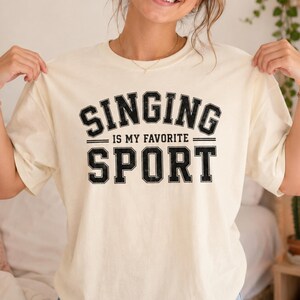 Op de afbeelding: Een crèmekleurig T-shirt met korte mouwen en zwarte, verweerde blokletters met de tekst SINGING IS MY FAVORITE SPORT. De tekst is verdeeld over drie regels, waarbij SINGING en SPORT in een groot lettertype staan en IS MY FAVORITE in een kleiner lettertype.