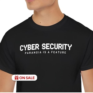 May include: A black t-shirt featuring white text that reads CYBER SECURITY in a pixelated font, with the words PARANOIA IS A FEATURE printed in a smaller, clean sans-serif font directly underneath the main heading on the chest area.