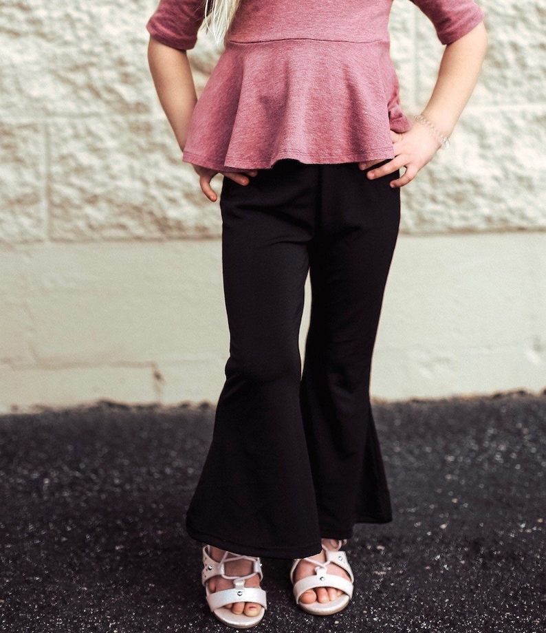 May include: A child wearing a mauve peplum top and black flared pants. The outfit is paired with white sandals. The background is a textured, off-white wall and dark pavement.