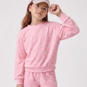 May include: A light pink, two-piece outfit featuring a crew neck sweatshirt and matching shorts. The set is adorned with a floral pattern. The outfit is paired with a light pink baseball cap.