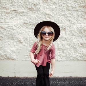 May include: A young person wearing a dark brown hat, sunglasses, a mauve top, black pants, and white sandals. The person is smiling and standing in front of a light-colored brick wall.