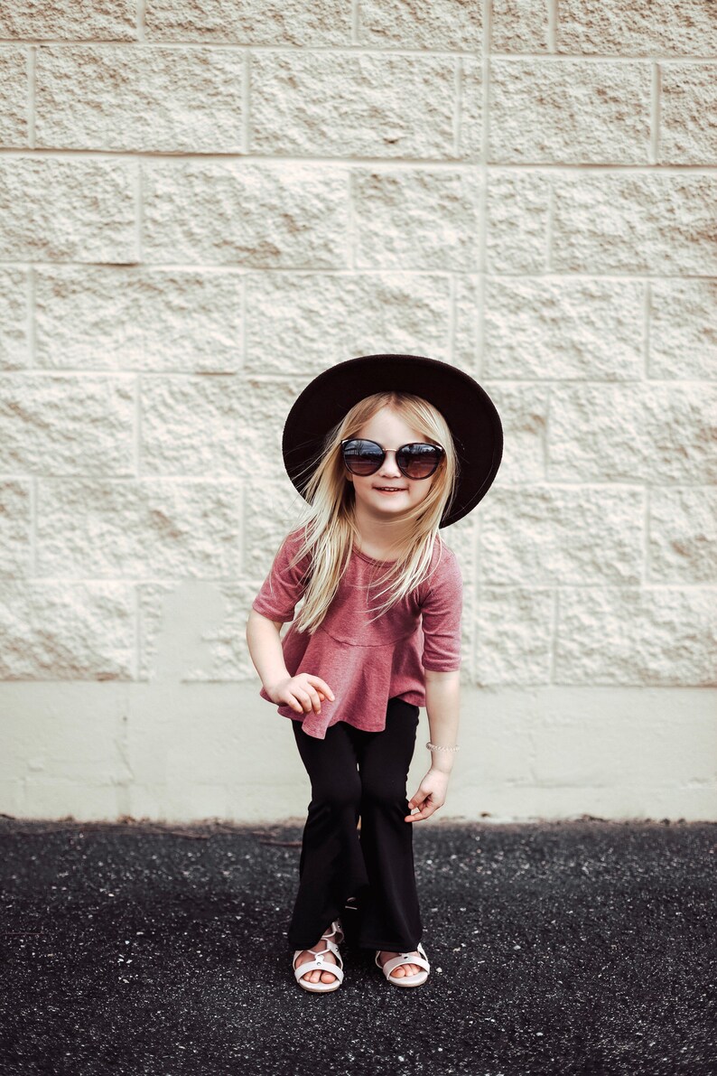May include: A young person wearing a dark brown hat, sunglasses, a mauve top, black pants, and white sandals. The person is smiling and standing in front of a light-colored brick wall.