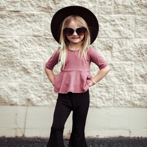 May include: A young person wearing a mauve top with a peplum waist, black bell-bottom pants, sunglasses, and a black hat. The outfit is paired with white sandals. The background is a textured, light-colored brick wall.