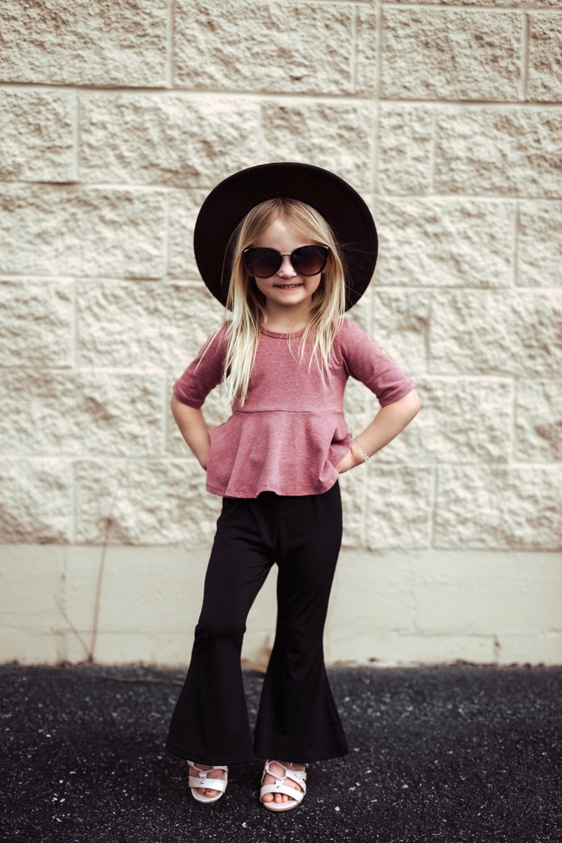 May include: A young person wearing a mauve top with a peplum waist, black bell-bottom pants, sunglasses, and a black hat. The outfit is paired with white sandals. The background is a textured, light-colored brick wall.