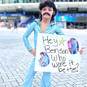May include: A person in a light blue jumpsuit with a matching shirt and white sneakers, holding a sign that says "Hey Benson Who wore it better?" The person is wearing a mustache and a wig. The background includes a building.