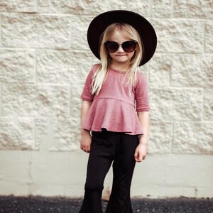 May include: A young person wearing a dark brown hat, sunglasses, a dusty rose top with a peplum, and black bell-bottom pants. The person is also wearing white sandals. The background is a light-colored brick wall.