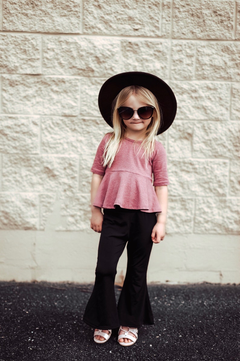 May include: A young person wearing a dark brown hat, sunglasses, a dusty rose top with a peplum, and black bell-bottom pants. The person is also wearing white sandals. The background is a light-colored brick wall.