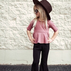 May include: A young person wearing a maroon top, black flared pants, sunglasses, and a brown hat. The outfit is paired with white sandals. The background is a textured, light-colored brick wall.