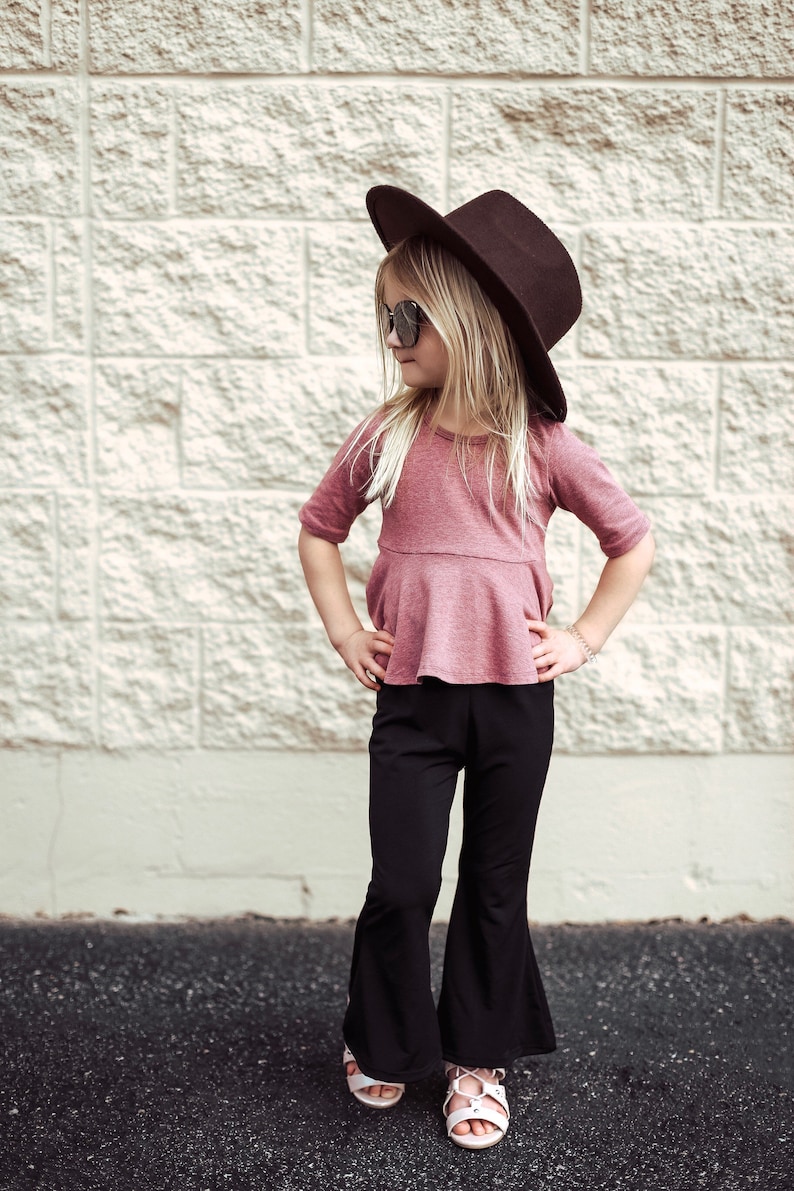 May include: A young person wearing a maroon top, black flared pants, sunglasses, and a brown hat. The outfit is paired with white sandals. The background is a textured, light-colored brick wall.