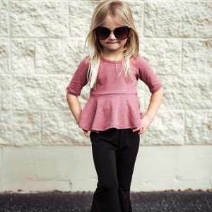 May include: A young girl wearing a mauve-colored top with a peplum waist and black flared pants. She is wearing sunglasses and sandals. The background is a textured, light-colored brick wall.