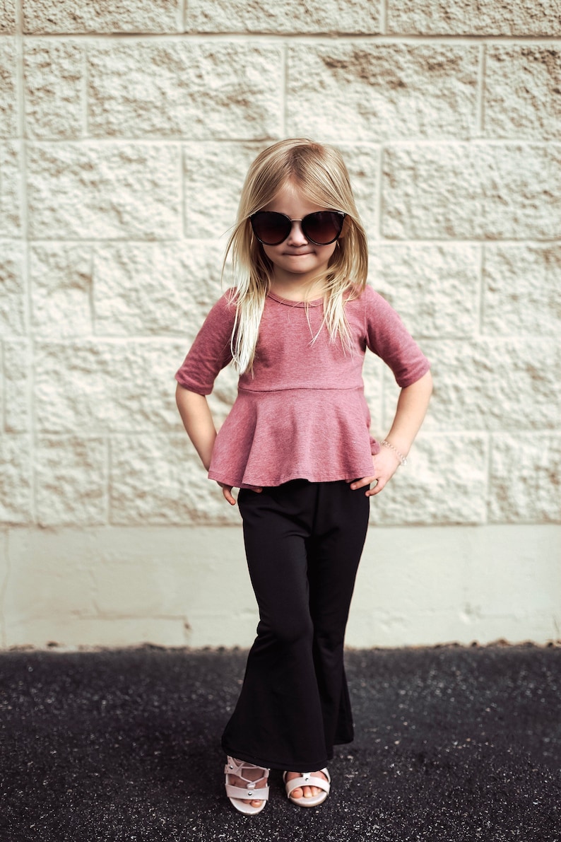 May include: A young girl wearing a mauve-colored top with a peplum waist and black flared pants. She is wearing sunglasses and sandals. The background is a textured, light-colored brick wall.