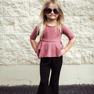 May include: A young person wearing a mauve-colored top with a peplum design, paired with black flared pants and sunglasses. The outfit is completed with white sandals. The background is a textured, light-colored brick wall.