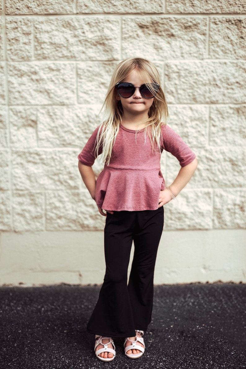 May include: A young person wearing a mauve-colored top with a peplum design, paired with black flared pants and sunglasses. The outfit is completed with white sandals. The background is a textured, light-colored brick wall.