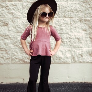 May include: A young person wearing a dark brown hat, sunglasses, a mauve top, and black bell-bottom pants. The outfit is completed with white sandals. The background is a light-colored brick wall.