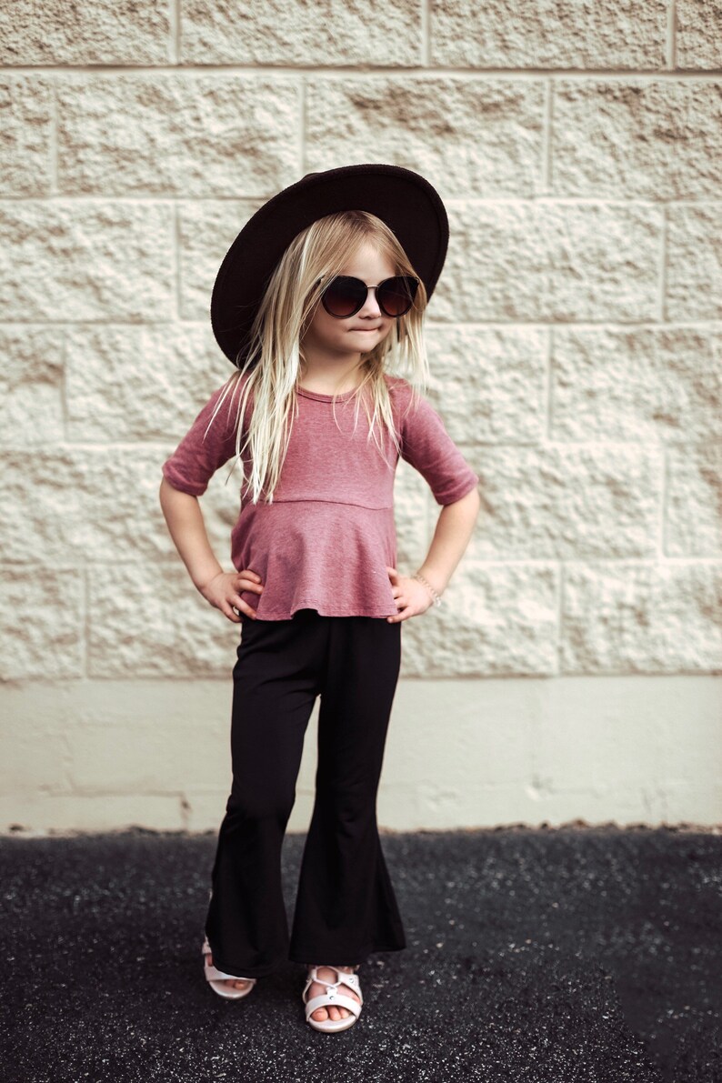 May include: A young person wearing a dark brown hat, sunglasses, a mauve top, and black bell-bottom pants. The outfit is completed with white sandals. The background is a light-colored brick wall.