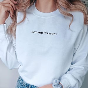 Könnte beinhalten: Ein weißes Sweatshirt mit dem Text "NOT FOR EVERYONE" in Schwarz. Das langärmlige Oberteil wird mit einer blauen Jeans kombiniert. Am Handgelenk wird ein hellblaues Scrunchie getragen.