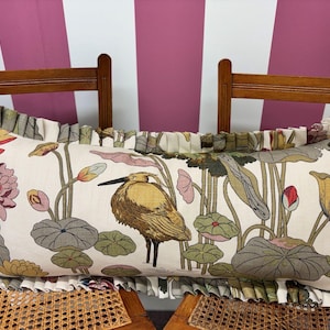 May include: A rectangular cushion with a botanical design. The cream-coloured fabric displays a detailed illustration of a bird, lotus flowers, and lily pads. The edges are trimmed with a ruffled border in complementary colours.