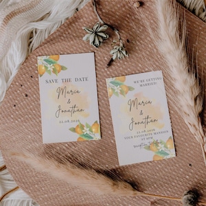 Puede incluir: Dos tarjetas de invitación de boda con ilustraciones florales y de cítricos. Las tarjetas dicen "Save the Date" y "We're Getting Married!" con los nombres "Maria & Jonathan" y la fecha 22.08.2026. Las tarjetas están sobre una superficie texturizada de tonos neutros.