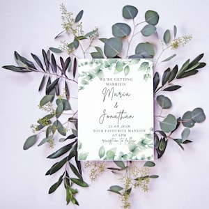 Puede incluir: Una invitación de boda con el texto "We're getting married! Maria & Jonathan 22.08.2026 Your favourite mansion at 6pm reception to follow." La invitación está rodeada de eucalipto verde y ramas de olivo sobre un fondo blanco.