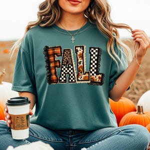 May include: A teal short-sleeved t-shirt featuring the word FALL printed in large letters. Each letter has a unique pattern including orange plaid, black and white checkerboard, leopard print, and floral designs, all outlined with black lace trim.