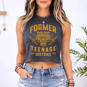 May include: A charcoal gray sleeveless crop top featuring a mustard yellow graphic print of a tiger head, lightning bolts, and the text "FORMER TEENAGE DIRTBAG" with "TRD" and "MRK" lettering, paired with light blue denim shorts.