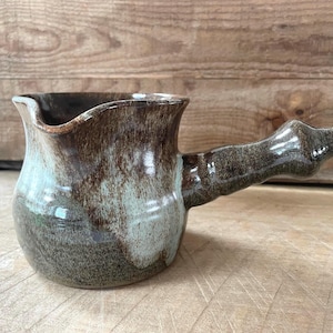 May include: A handcrafted ceramic jug with a handle. The jug features a brown and green glaze, with a distinctive form and a pouring spout. The handle is robust and solid. The jug is probably used for serving drinks.
