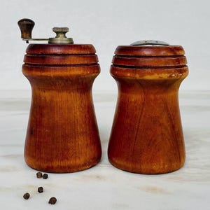 Peut inclure: Une paire de moulins à sel et à poivre en bois vintage. Le moulin à sel a une manivelle en métal et le poivrier a un dessus en métal avec des trous. Les deux sont en bois de couleur chaude et reposent sur une surface claire.