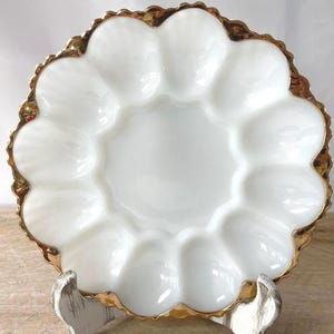 Pode incluir: Um prato redondo para ovos em vidro leitoso branco com doze cavidades ovais individuais dispostas num padrão circular. A borda recortada do prato tem um acabamento com um friso dourado metálico que segue as curvas do rebordo.