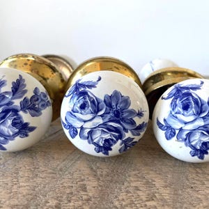 May include: Three round door knobs featuring a white ceramic surface decorated with a blue floral pattern. Each knob has a polished gold-tone metal base. The floral design consists of detailed blue roses and leaves arranged across the front.
