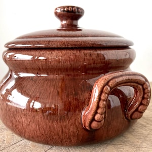 May include: A round, glazed ceramic pot in a deep reddish-brown color with a matching lid. The pot features two side handles with a textured, beaded design and a small, round knob on the center of the lid, which also has a beaded edge.