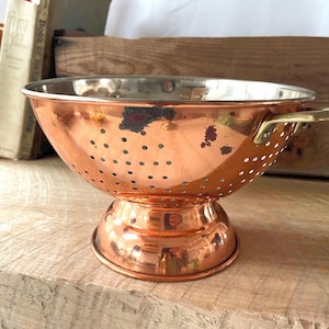 May include: A copper colander with a shiny surface and small holes. It has a pedestal base and brass handles. The colander is on a wooden surface, with books and a wooden box in the background.