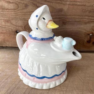 Könnte beinhalten: Eine Keramik-Teekanne in Form einer Gans, die eine weiße Haube mit blauen Punkten trägt. Die Gans hat einen gelben Schnabel und ist mit einer weißen Schürze mit rosa und blauem Saum bekleidet und hält ein kleines blaues Kännchen auf einem Tablett.