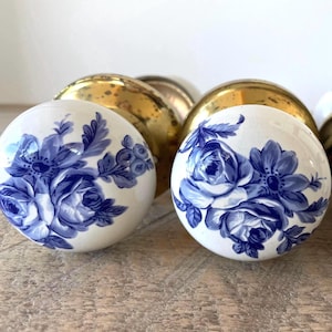 Peut inclure: Deux poignées de porte rondes dotées d'une surface en céramique blanche avec un motif floral bleu. Chaque poignée est fixée sur une base en laiton poli présentant des signes d'usure et de patine. Le motif floral représente des roses détaillées.