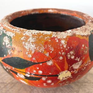 May include: A round, bowl-shaped ceramic vessel featuring a weathered, textured surface with a hand-painted floral design. The exterior displays vibrant red and orange flower petals accented by dark green leaves, all overlaid with a distressed, white, crusty finish.