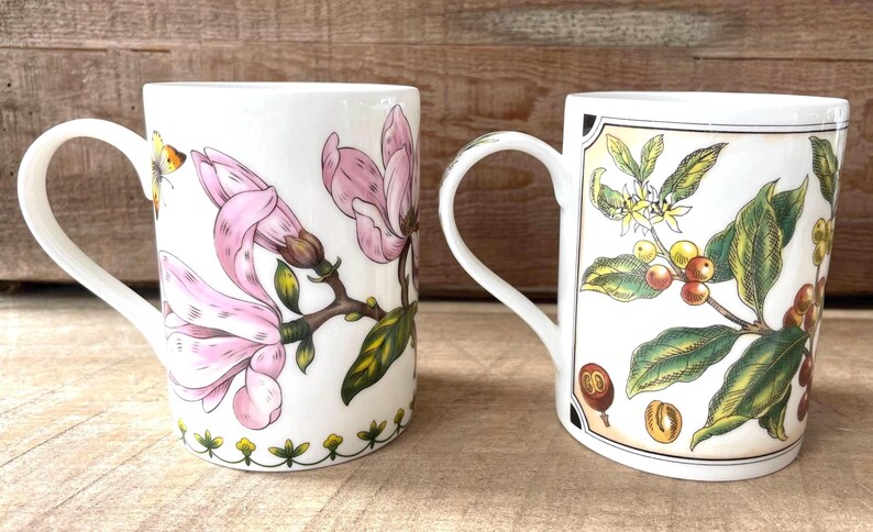 May include: Two white ceramic mugs featuring botanical illustrations. The left mug displays pink magnolia flowers and a small butterfly. The right mug features a framed illustration of coffee plant branches with green leaves, yellow flowers, and red coffee cherries.
