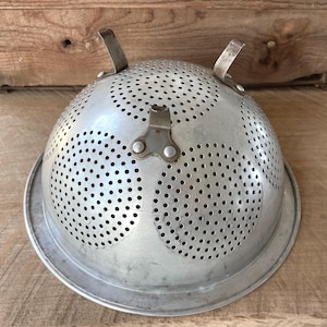 Puede incluir: Un colador vintage de color plateado con numerosos agujeros pequeños para escurrir. Cuenta con dos asas y una base robusta. El colador es de metal y está diseñado para uso en la cocina.