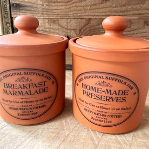 May include: Two terracotta-coloured ceramic jars with lids. One jar reads "Breakfast Marmalade" and the other reads "Home-Made Preserves." Both jars have a black oval label with text. The jars are labelled "The Original Suffolk Jar" and "Henry Watson Pottery."