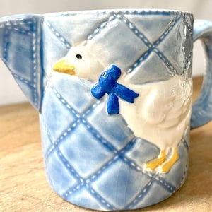 May include: A ceramic jug featuring a duck design. The jug is light blue with a quilted pattern and a white duck with a blue bow. The duck has a yellow beak and feet. The jug has a handle and a spout.