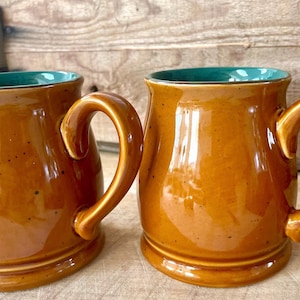 Puede incluir: Dos tazas de cerámica marrón con un acabado brillante e interiores color turquesa. Las tazas tienen asas grandes y curvas y están sobre una superficie de madera. Las tazas son probablemente para café o té.