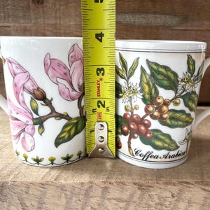 May include: Two white ceramic mugs positioned next to a yellow Stanley measuring tape showing a height of approximately 4.5 inches. The left mug features pink magnolia flowers, while the right mug displays coffee plant illustrations with the text Coffea Arabica.