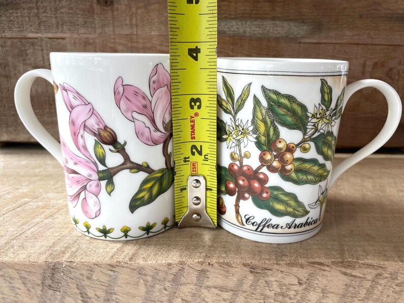 May include: Two white ceramic mugs positioned next to a yellow Stanley measuring tape showing a height of approximately 4.5 inches. The left mug features pink magnolia flowers, while the right mug displays coffee plant illustrations with the text Coffea Arabica.