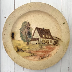May include: A circular, beige ceramic plate featuring a hand-painted rural scene. The image shows a white house with a brown roof, a tree, and a path. The plate has a rustic, textured finish and is signed by the artist.