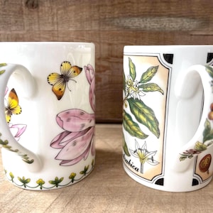 May include: Two white ceramic mugs featuring botanical illustrations. The left mug displays pink flowers and yellow butterflies, with a green vine pattern on the handle. The right mug shows coffee plant leaves, white flowers, and the text Arabica.