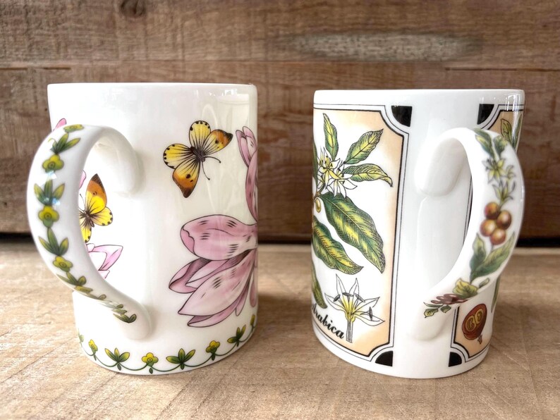 May include: Two white ceramic mugs featuring botanical illustrations. The left mug displays pink flowers and yellow butterflies, with a green vine pattern on the handle. The right mug shows coffee plant leaves, white flowers, and the text Arabica.